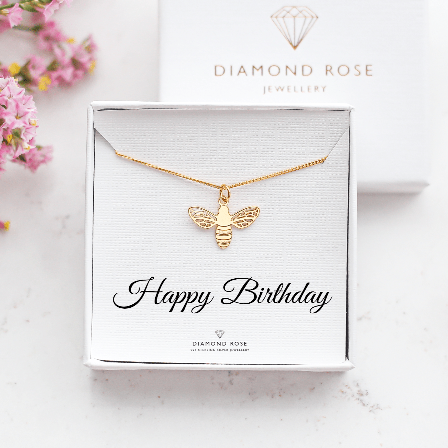Gold bee necklace in a 'Happy Birthday' card with Diamond Rose Jewellery branding, surrounded by pink flowers.