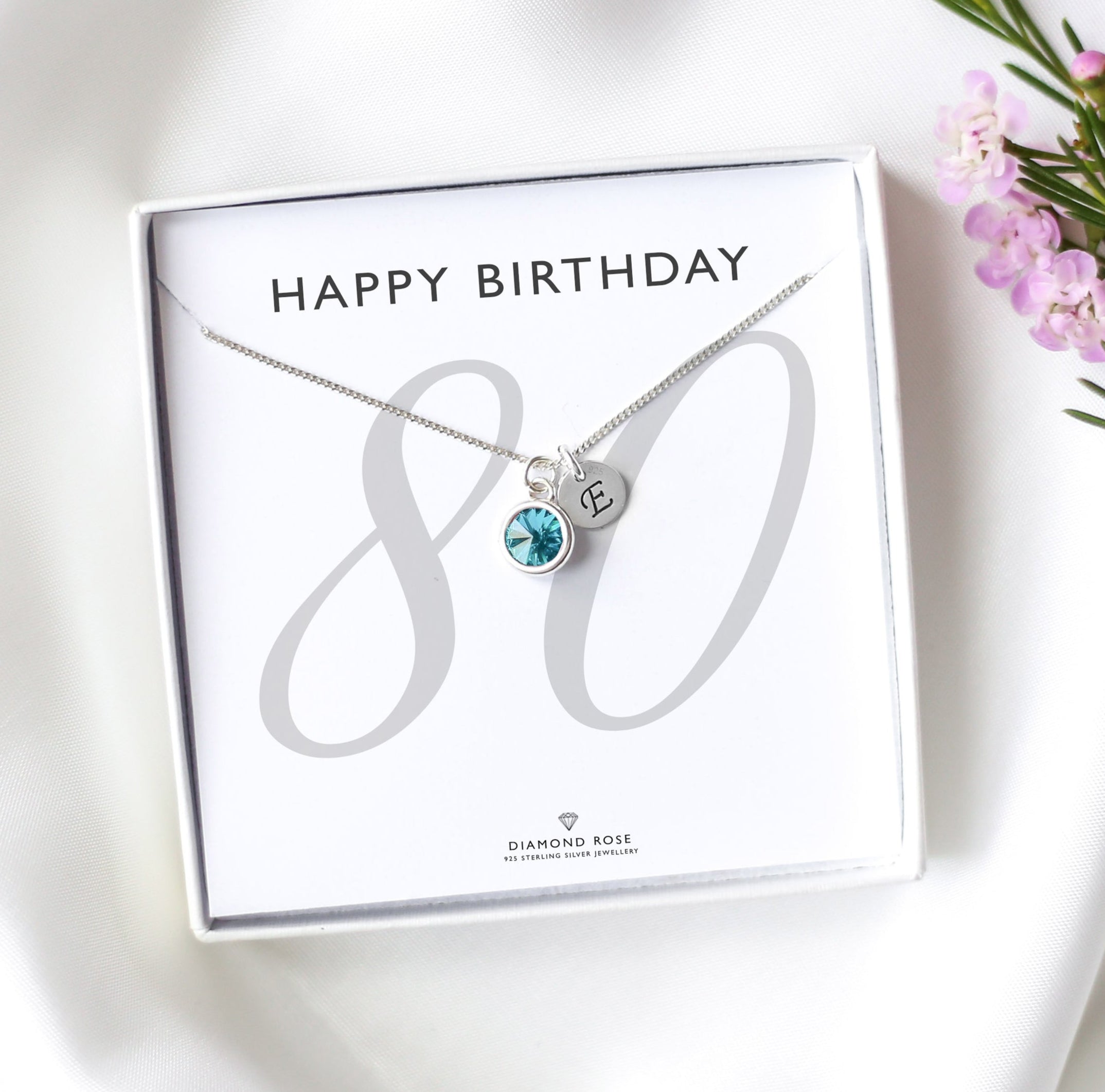 Silver necklace with a birthstone in a 'Happy Birthday' box on a white surface with flowers.