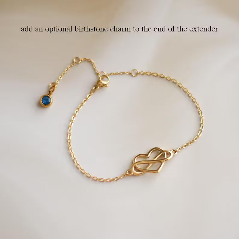 A delicate gold chain bracelet with a heart-shaped infinity link centerpiece, displayed on a light cream background. The text at the top reads 'add an optional birthstone charm to the end of the extender', pointing to a small blue gemstone charm on the left side of the chain.