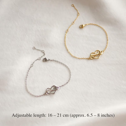 Two delicate chain bracelets with interlocking heart charms, one in gold and one in silver, displayed on a textured white fabric background with text indicating adjustable length 16-21 cm.
